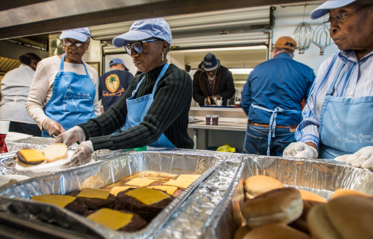 Volunteers in matching aprons serving a hot community meal at a soup kitchen buffet line.