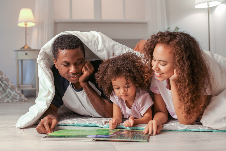 A father, mother, and young daughter smiling together while reading a book under a blanket at home
