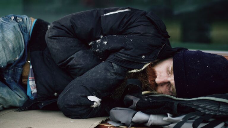 A homeless man sleeping outdoors on cardboard during winter in a city.